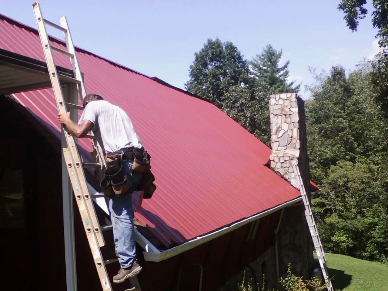Roofer on a ladder beside a red metal roof and stone chimney — Soffit & Fascia Repair by Roofing Brothers in Browerville